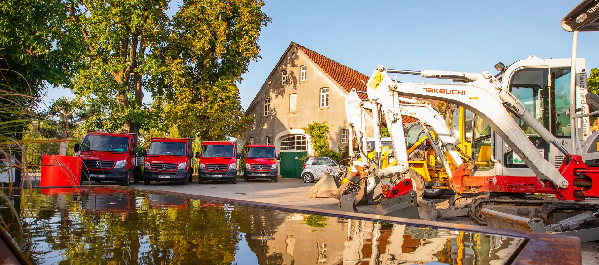 Baumaschinen und rote Fahrzeuge auf einem Hof, mit Spiegelung in einer Wasserfläche.